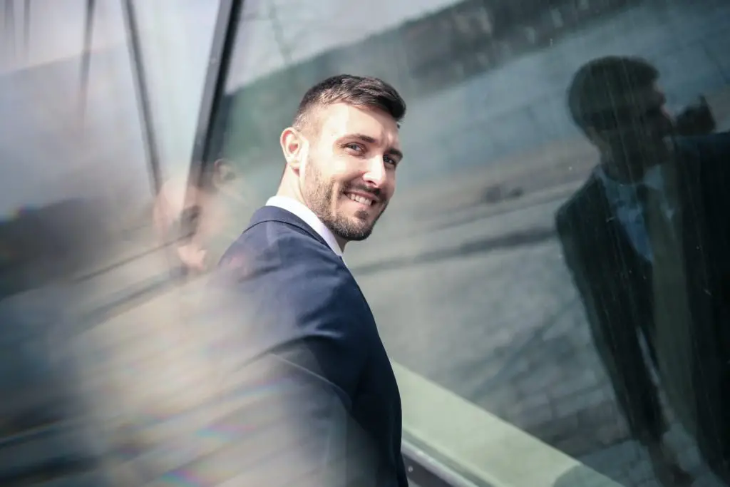 A young businessman in a suit stnding in front of a glass building wall ands looking back at the camera smiling. ﻿is MBA necessary to start a business