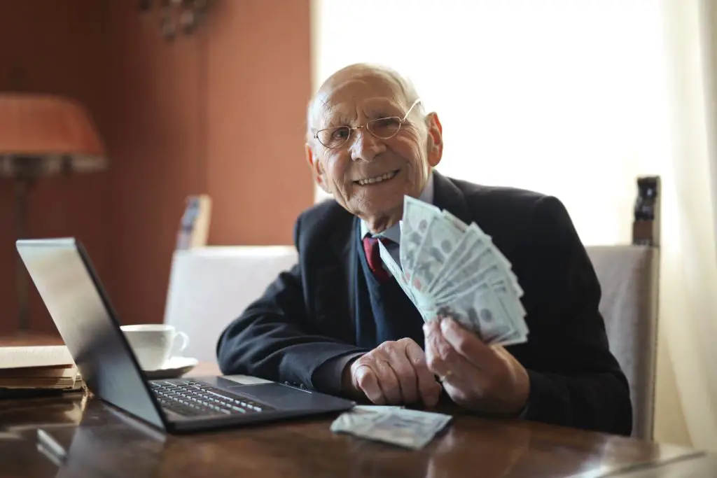 A mature male entrepreneur in a suit sitting in front of his open laptop on his desk and holding a unch of ddollar bills in his hand
