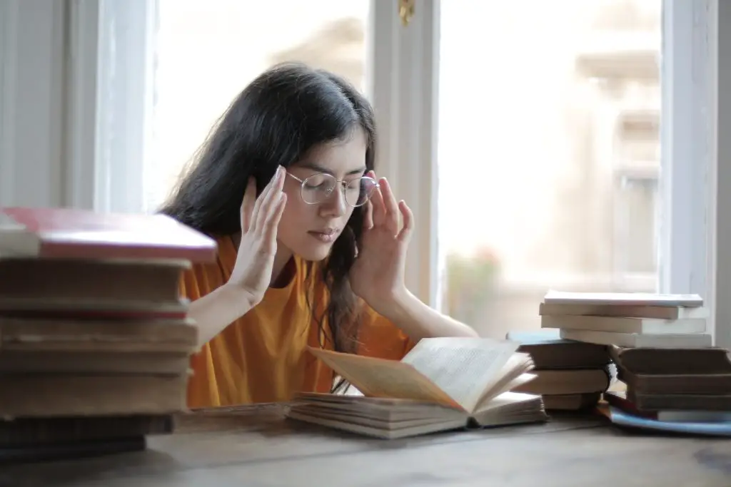 A female brunette law school graduate in glasses sitting by the table preparing for hard bar exam with many books around.