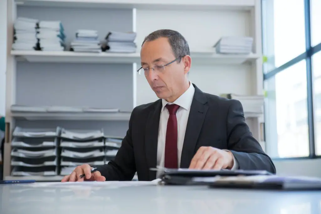 Male accountant in suit in the office behind the desk looking through paperwork